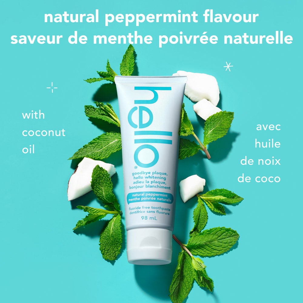 A tube of goodbye plaque hello whitening fluoride free toothpaste stands upright surrounded by fresh mint leaves and coconut pieces on a turquoise background. Text describes &quot;natural peppermint flavour&quot; in English and French, and &quot;with coconut oil&quot; in both languages.