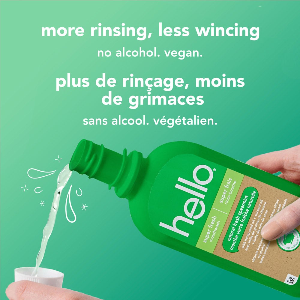 A hand pours hello brand green super fresh mouthwash into the mouthwash bottle cap with a light green background. The bottle label is clear. White text reads, &quot;more rinsing, less wincing – no alcohol. vegan.&quot; with French translation below.