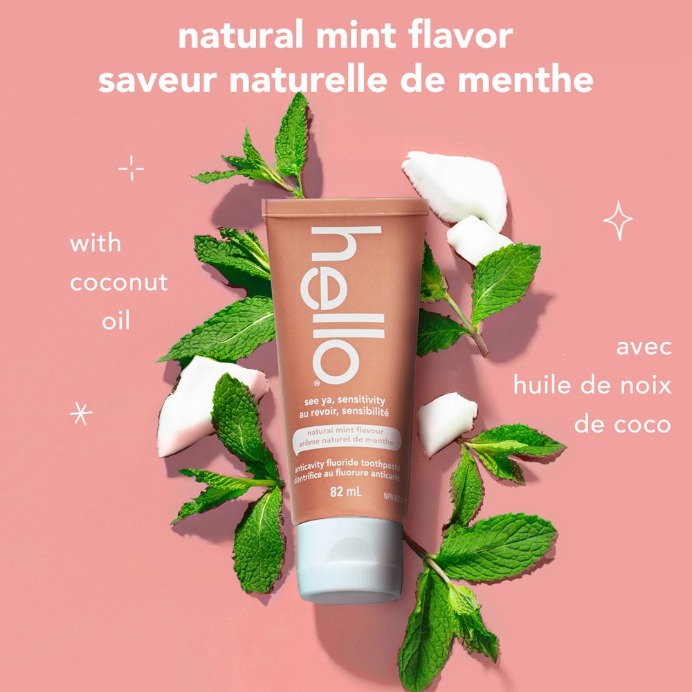 A tube of hello See Ya Sensitivity toothpaste is surrounded by fresh mint leaves and pieces of coconut on a pink background. Text highlights &quot;natural mint flavor&quot; and &quot;with coconut oil&quot; in both English and French.