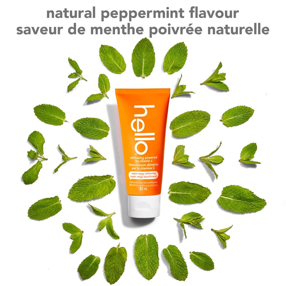 An orange tube of hello super mega whitening toothpaste sits upright in the center, surrounded by fresh peppermint leaves arranged in a circle on a white background. Text at the top says &quot;natural peppermint flavour&quot; in English and French.