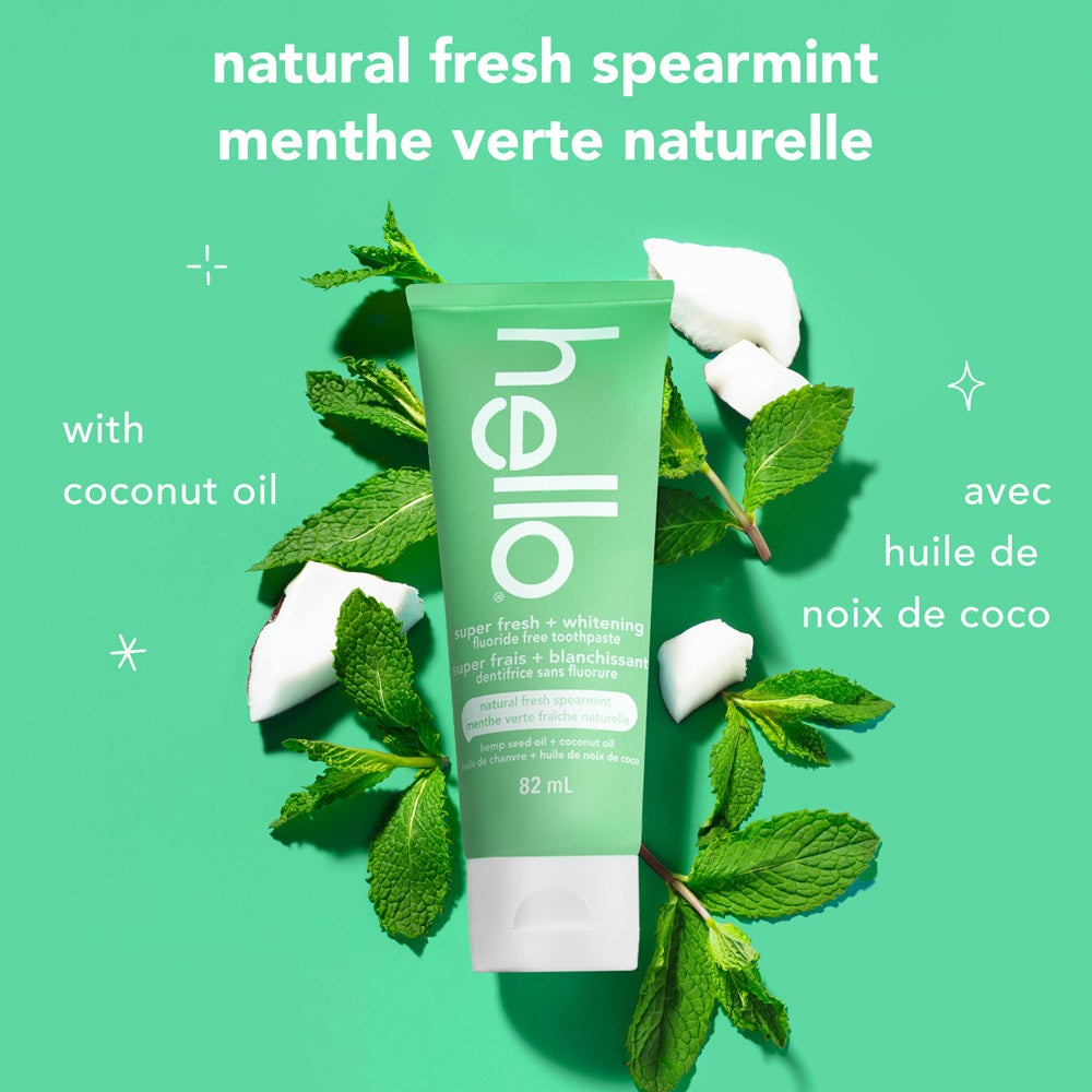 A green tube of hello super fresh + whitening fluoride free toothpaste stands at the center on a solid green background, surrounded by fresh mint sprigs and coconut pieces. Above the product, the text reads: &quot;natural fresh spearmint.&quot; To the left, it says &quot;with coconut oil.&quot;