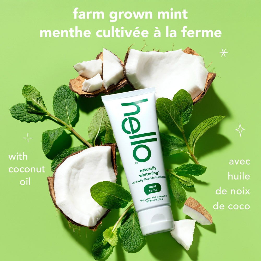 A tube of hello naturally whitening fluoride toothpaste with farm grown mint stands upright among fresh mint leaves and cracked coconut pieces on a light green background. Text above the tube says, &quot;farm grown mint&quot; in English and French, and &quot;with coconut oil&quot; in both languages.