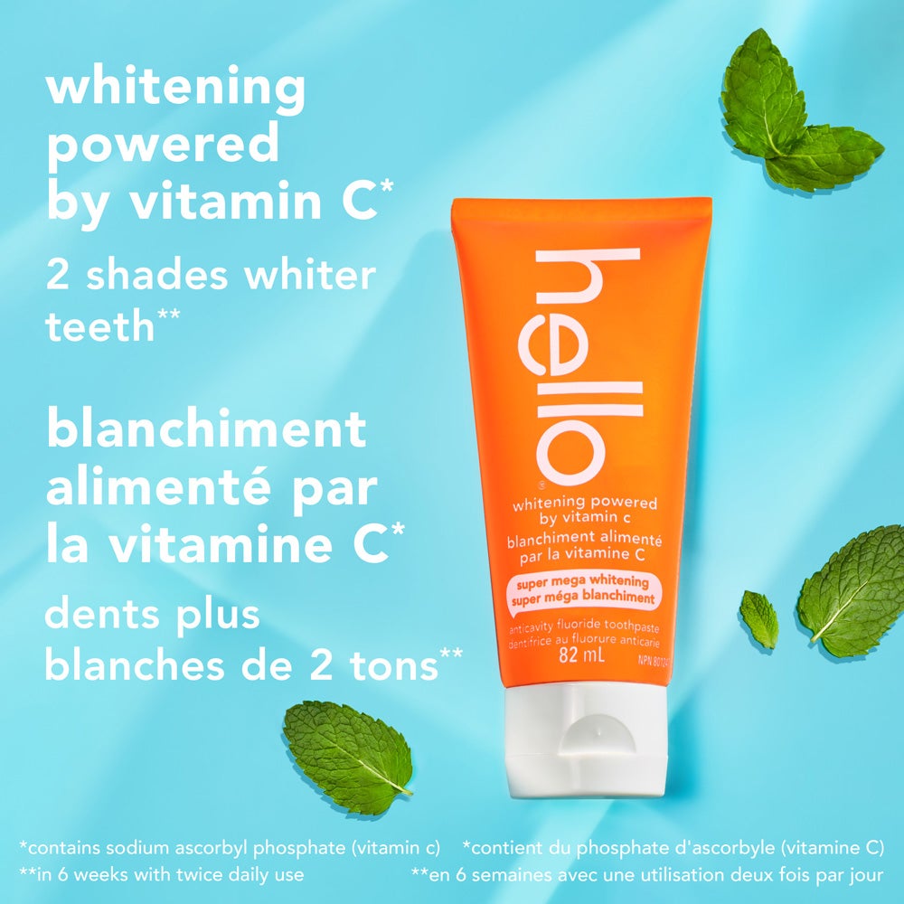 An orange tube of hello super mega whitening toothpaste stands upright on a light blue background scattered with fresh mint leaves. Text highlights &quot;whitening powered by vitamin C&quot; and &quot;2 shades whiter teeth&quot; in English and French, with additional small print disclaimers at the bottom.
