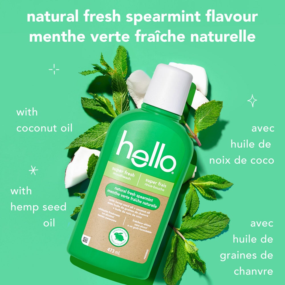 A green bottle of hello super fresh mouthwash is placed on a mint-colored background surrounded by fresh mint leaves, coconut pieces, and hemp seeds. Text says, &quot;natural fresh spearmint flavour&quot; in English and French, with smaller text indicating &quot;with coconut oil&quot; and &quot;with hemp seed oil&quot; in both languages.