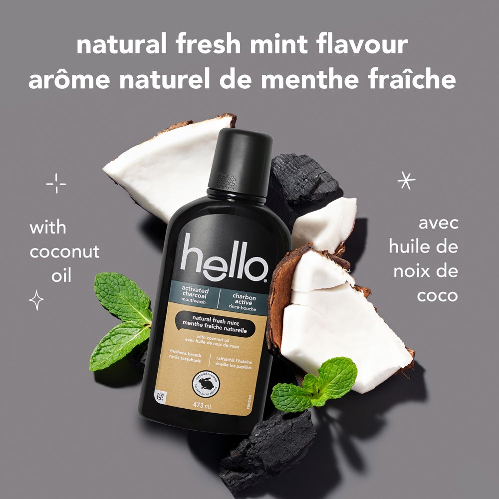 A black bottle of hello activated charcoal mouthwash is centered on a light grey background, surrounded by pieces of coconut, black charcoal chunks, and fresh mint sprigs. The text above reads: &quot;natural fresh mint flavour.&quot; To the left, it says &quot;with coconut oil.&quot;