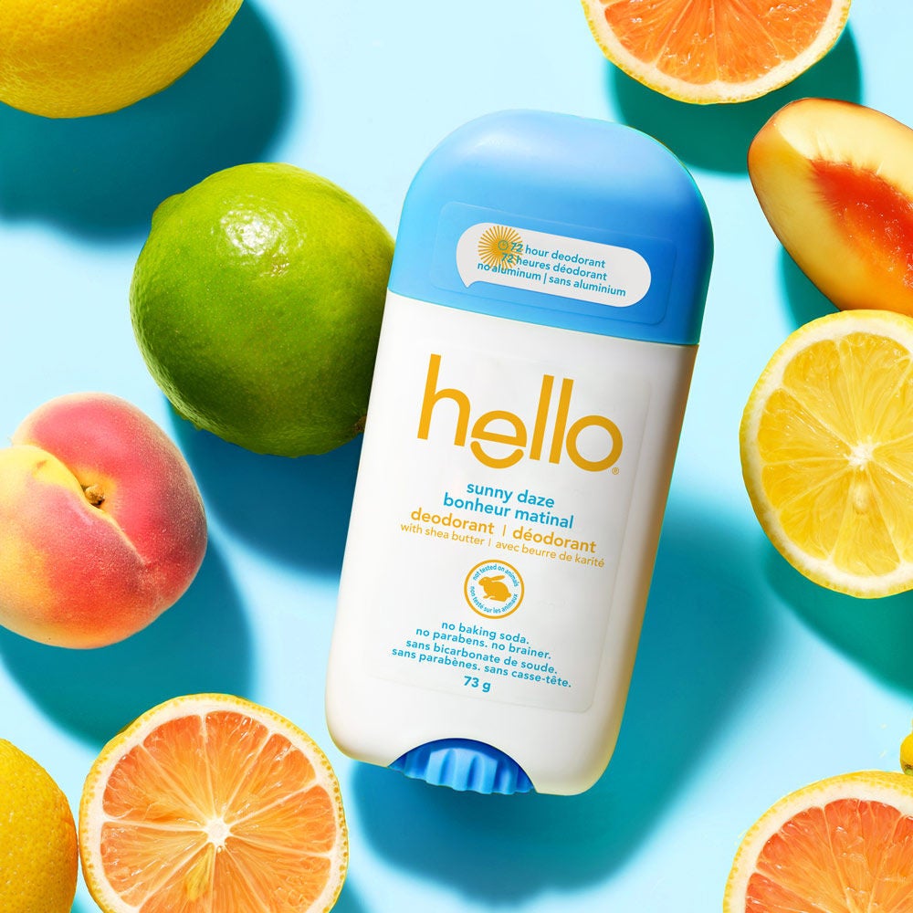 Hello Sunny Daze 72-hour deodorant stick centered on a sky-blue surface surrounded by fresh citrus fruit slices including lemons, limes, peaches, and grapefruits